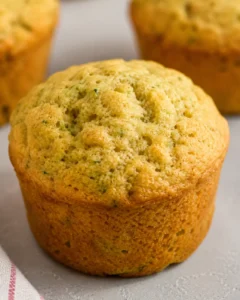 Close-up of moist, golden zucchini muffin with background muffins on a white surface.